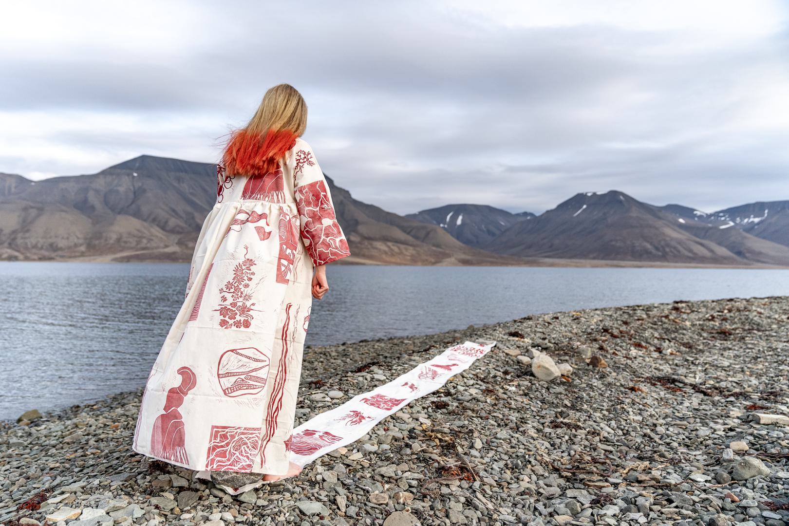 En dame kledd i en lang kjole som er hvit med rødt mønster fra samisk kultur. Hun står ved en fjord i arktisk landskap
